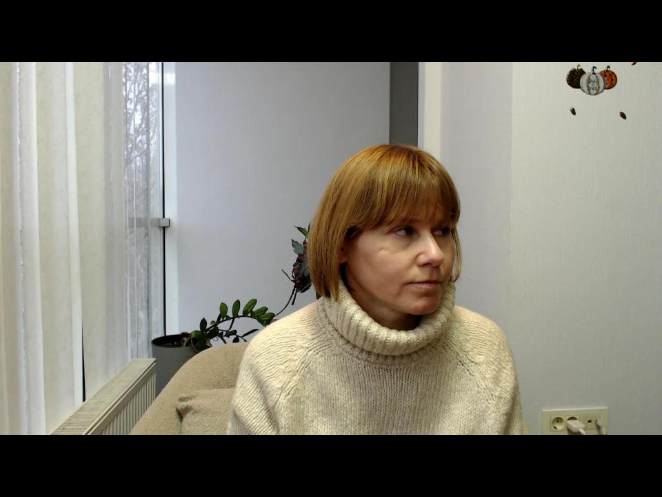 Marlenda with short orange hair, wearing a fluffy white turtleneck, sits in a room. She has a small beauty mark under her right eye and a slight smile.