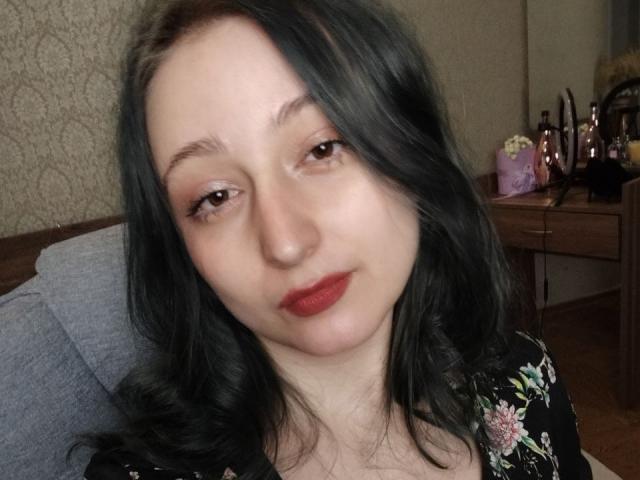 TwinkyKiss has pale skin, dark wavy hair, red lipstick, and brown eyes, wearing a black floral top, sitting with a slight smile, looking relaxed.