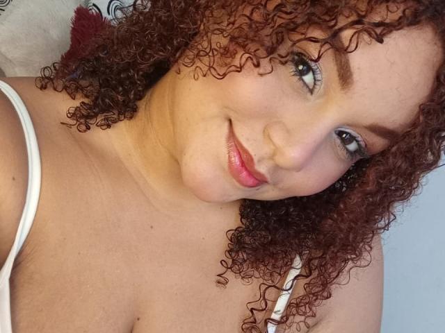 YeraCarey with curly red hair, smiling, wearing a white tank top, showing her tits and a cute face with pink lips and blue eyes.