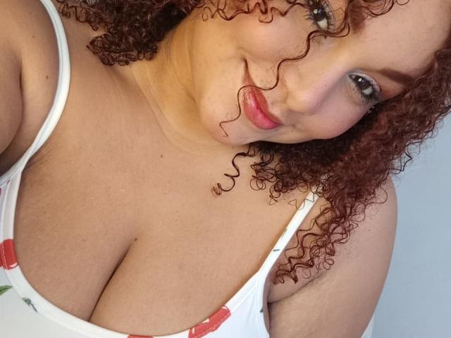 YeraCarey has curly red hair, fair skin, and large tits in a white tank top with red accents, smiling with pink lips and green eyes in a selfie.