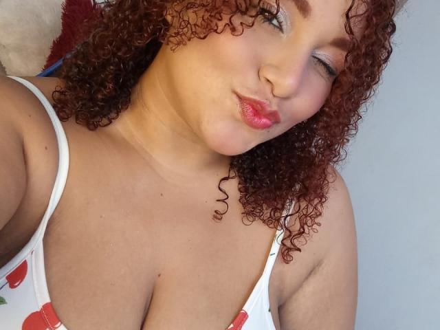 YeraCarey with curly red hair, pink lipstick, and white tank top showing big tits, puckering lips in a selfie. Light skin, one eye closed, subtle pink eye shadow.