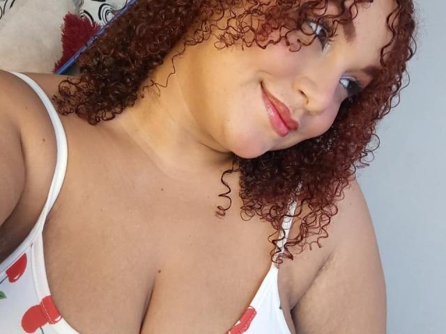 YeraCarey has curly red hair, light skin, and is smiling at the camera with pink lips. She's wearing a white tank top showing her big tits.