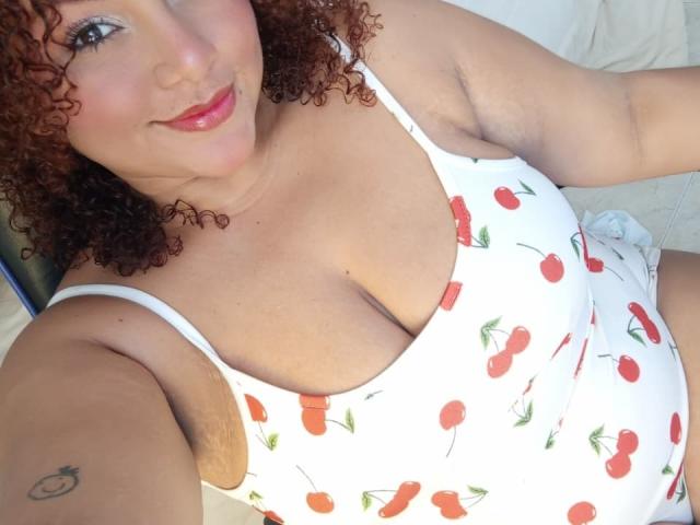 YeraCarey with curly red hair, smiling, wearing a white cherry print tank top, showing her big boobs, tattoo on right arm, lying down, white background.