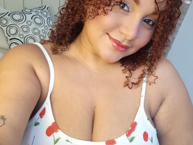 Curly red-haired YeraCarey with a smile, wearing a white dress with red flowers, showing her large, round boobs. Her light skin and pink lips are visible.