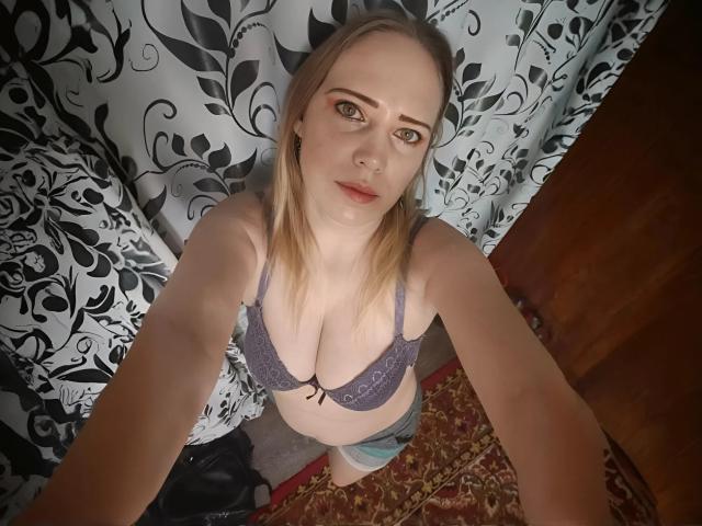 JenniDeep, avec ses cheveux blonds, portant un soutien-gorge en dentelle violet, montrant ses seins, debout dans une pièce avec des rideaux floraux noirs et blancs, regardant vers le haut de la caméra..