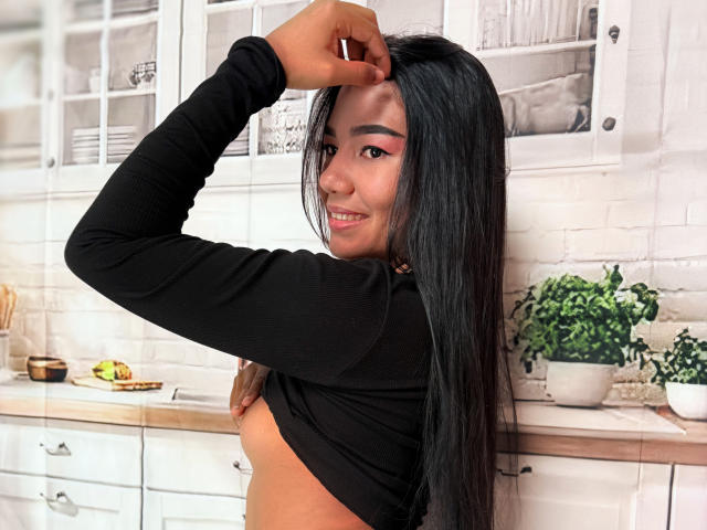 AnaPorto, com cabelos longos e pretos, vestindo uma camisa preta de manga comprida que revela seus pequenos seios, levantando o braço e sorrindo em uma cozinha branca. Plantas em vasos e utensílios de cozinha ao fundo..