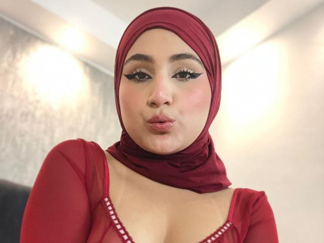 HiilaryDolce with a red headscarf, wearing a red top with white dots, pouting her full lips, has black eyeliner, and a light skin tone, showing her medium-sized tits.