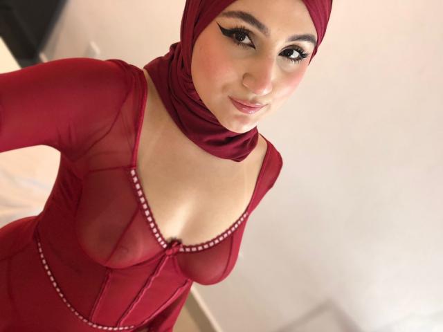 HiilaryDolce wearing a red sheer dress with visible tits, red headscarf, and bold eyeliner, smiling at the camera. She's got a cute face and a sexy look.