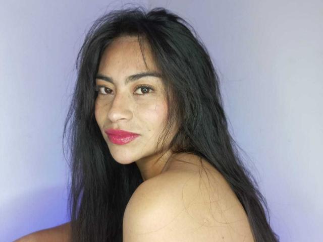 Emily Mitchell tem cabelos longos, pretos e levemente desgrenhados, olhos castanhos e batom rosa, olhando por cima do ombro com um leve sorriso, mostrando seus ombros nus..