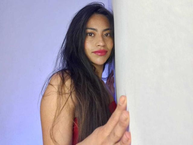 Emily Mitchell, avec ses longs cheveux noirs, son rouge à lèvres rouge et son haut rouge, apparaissant derrière un pilier blanc, tenant le pilier avec sa main droite..