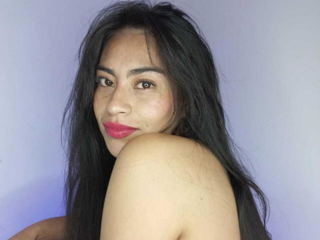 Emily Mitchell, com cabelos longos e pretos, olhos castanhos, batom vermelho e ombros descobertos, olhando por cima do ombro com um leve sorriso. Seus seios estão parcialmente visíveis..