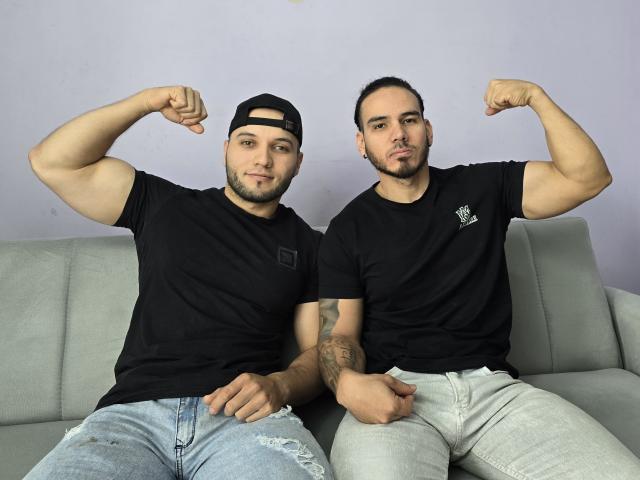 Two muscular SteveJhustins with flexed biceps, black t-shirts, and jeans, sitting on a gray couch, one with a black cap and the other with tattoos.