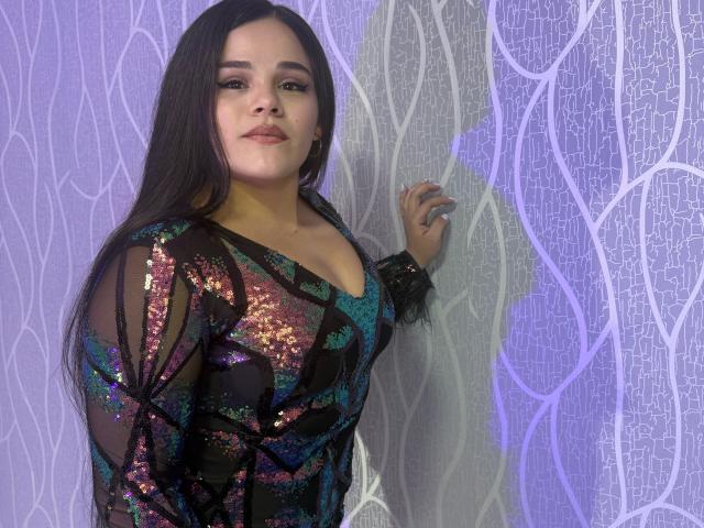 PammellaStone with long dark hair, wearing a sheer black dress with colorful sequins, standing against a textured wall, looking at the camera with a slight smile.