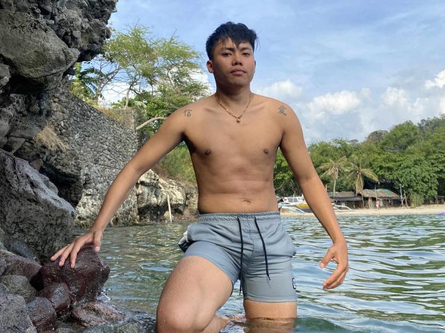 Rayzen69, shirtless, with tattoos on both shoulders, wearing gray shorts, standing in shallow water, hands on rocky shore, looking at camera, short black hair, medium build, tan skin.