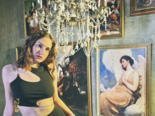StellaMari with wavy brown hair, wearing a black sports bra, standing in front of paintings, including a classical nude with wings. Chandelier above, crystal drops, glowing.