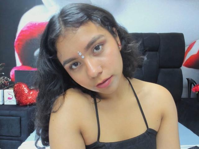 JuliaBennett with wavy black hair, brown skin, and small nose stud, wearing a black halter top, looking at the camera with a slight pout. Her tits are small.