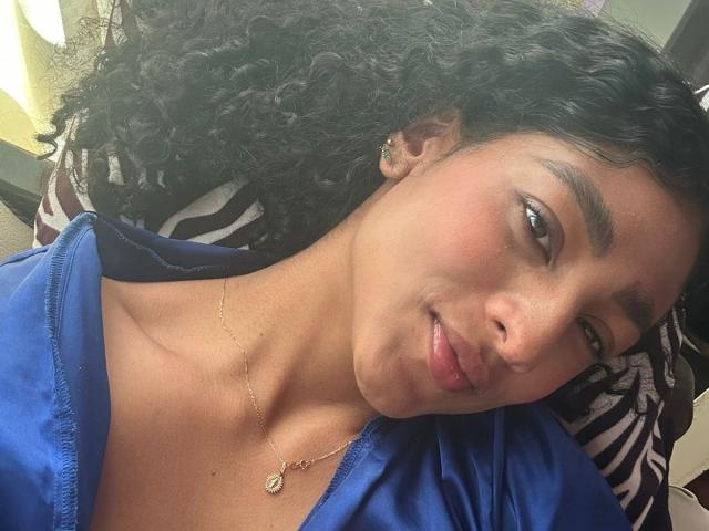NatalyBlonder with curly black hair, medium brown skin, and subtle makeup, wearing a blue shirt and gold necklace, lying down with a relaxed, slightly pouty expression.