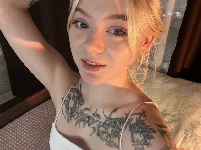 EnchantingEyes has blonde hair, blue eyes, and detailed black tattoos covering her chest. She's wearing a white tank top, smiling, and raising her arm. Her tits are partially visible.