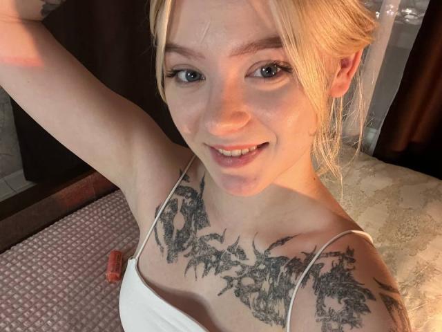EnchantingEyes with blonde hair, blue eyes, and tattoos on her chest and shoulders, smiling at the camera, wearing a white tank top, arm raised.