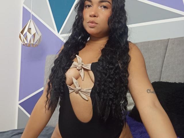 Alice Smity, con cabello negro, largo y rizado, vestida con un traje de baño negro de una sola pieza con detalles beige en forma de nudo, mostrando sus pechos a través del corte. Está sentada en una cama con un tatuaje..