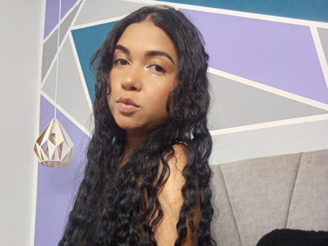 AliceSmity has long, wavy black hair, brown eyes, and full lips with a slight pout. She's looking at the camera, sitting with a gray headrest behind her.