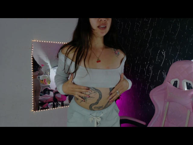 AsukaMisho with long brown hair, gray off-shoulder top, showing her bare tits, light gray pants, tattooed belly, hands on her belly, pink gaming chair, black wall.