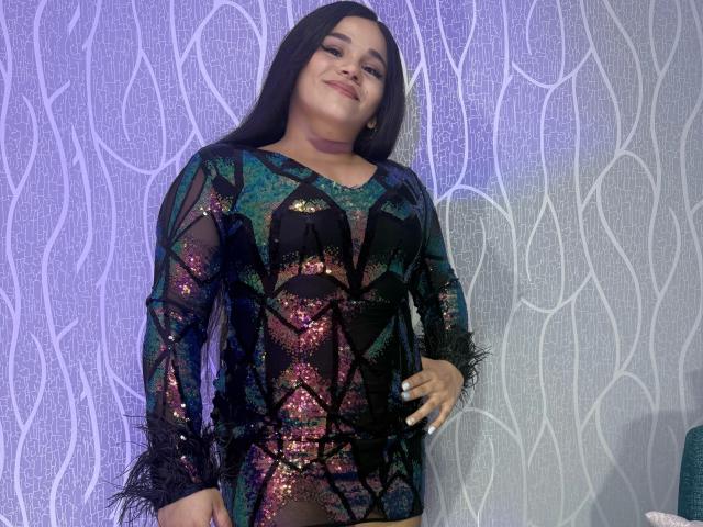 PammellaStone with long black hair, smiling, wearing a sheer, glittery black and green dress, exposing her small tits and black bra, standing against a purple, patterned wall.