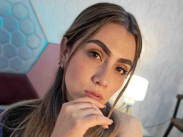 AriaRous with brown eyes, full eyebrows, and long brown hair, touching her slightly parted lips with her finger, showing a thoughtful expression. Her pink lips are slightly glossy.