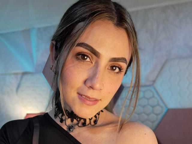 AriaRous has a cute face, brown eyes, and light skin. She's wearing a black choker with beads, and her brown hair is in a loose ponytail. Boobs not visible.