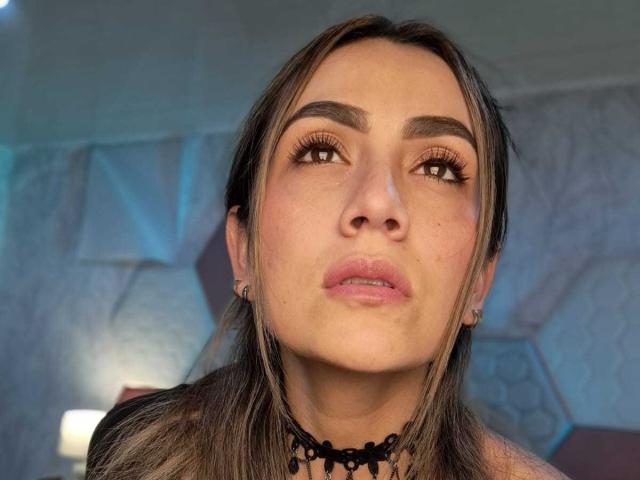 AriaRous has brown eyes, full pink lips, and a black lace choker, with long brown hair, subtle makeup, and small earrings, looking upward with a slightly pouty expression.