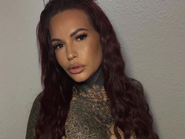 Aileenink-hot with dark wavy hair, tattoos covering her chest, nose ring, and full lips, staring into the camera with serious eyes.