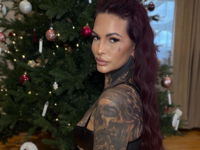 Aileenink-hot with dark red hair, tattoos covering her right arm, staring at camera, standing in front of decorated Christmas tree, wearing black lace top, looking sexy.