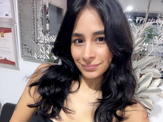 LyraRaven has long, wavy black hair, light brown skin, and a slight smile. She's wearing a black top, showing her small tits, in a well-lit room.