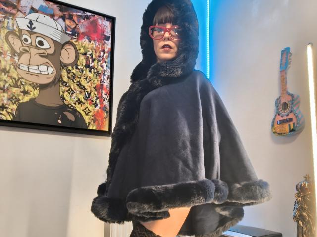 OrianaLaFrancaise in black fur cape, red glasses, and hood, standing with a slight turn, showing her bare ass, in a room with colorful art and a guitar.