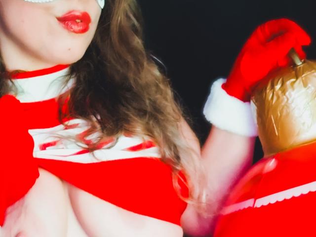 JuliaFrancaise with red lipstick, wavy brown hair, and a Santa outfit, exposing her big tits, holding a Santa hat, and wearing a white mask.