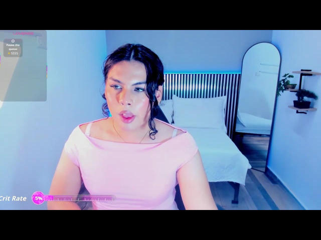 HottieAlii has medium-length dark hair in a ponytail, wearing a pink off-shoulder top. She has a slightly pouty expression, with pink lipstick. The room has a white bed with a striped headboard, a mirrored wall, and wooden shelves. The photo has a watermark in the bottom left corner that reads "Cutie Rate" with a pink logo. The image is brightly lit with a blue tint. HottieAlii's skin tone is light, and she has a small tattoo on her right arm.