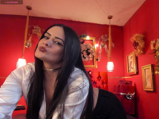 AnissaAli with long black hair, pouty lips, white shirt, black choker, leaning forward, red room, two hanging lights, gold decorations, and framed pictures, looking seductive.