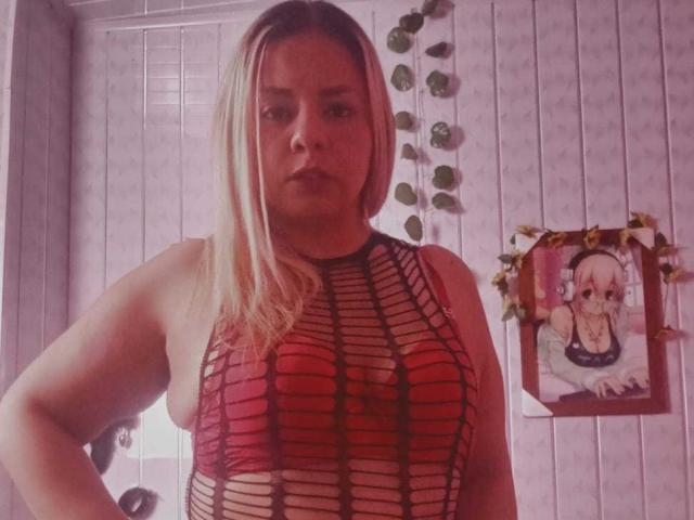 KathaleyaGrayyAss, con cabello rubio, vestida con una blusa de malla roja, negra y rosa que deja ver sus pechos, de pie en una habitación, con una expresión seria, y un lindo póster de anime de fondo..