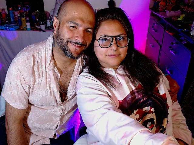 SektethLilith with bald head and beard, wearing a white patterned shirt, stands next to SektethLilith with glasses, black hair, and a white Marvel shirt, in a cozy setting.