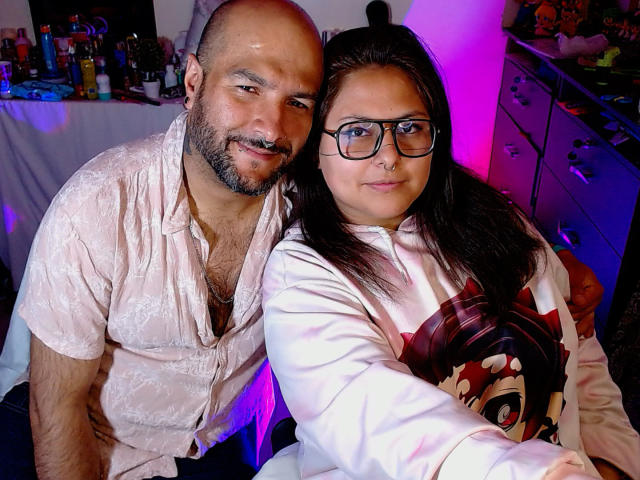 SektethLilith, een kale, baardgroeiige man met een gemiddeld postuur en lichte huid, gekleed in een roze shirt, zit naast SektethLilith, een vrouw met donker haar, een bril en een wit shirt..