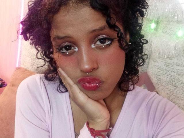 LunaKeller met krullend zwart haar, roze lippenstift en make-up, die een frons trekt met haar hand op haar wang, gekleed in een roze top, met een neusring en eyeliner, en die naar de camera kijkt..