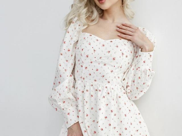 GlariyaHot with blonde hair wearing a white floral dress, puffy sleeves, and a scoop neckline, hand on chest, soft skin, subtle smile, white background, cute and feminine.
