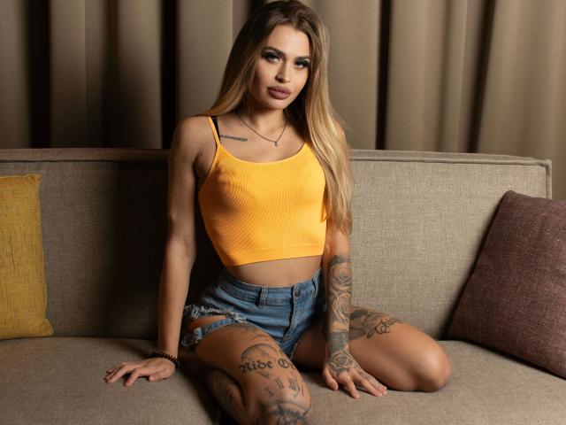 Simsibaby-hot with long blonde hair, yellow crop top, and denim shorts, sitting on a couch, showing tattoos on thighs, and a confident smile, with a yellow and brown pillow.