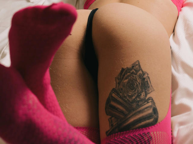 Simsibaby-hot's ass in black panties, pink socks, rose tattoo on right thigh, lying on white bed. Tattoo is detailed, realistic rose with ribbon. Socks are slightly fuzzy.