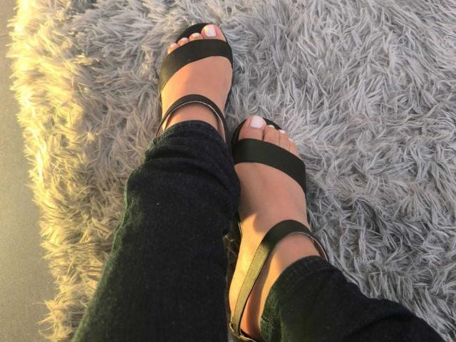Los pies de Heyly en sandalias negras de tiras descansan sobre una alfombra gris, la luz del sol arroja un cálido resplandor en sus dedos.