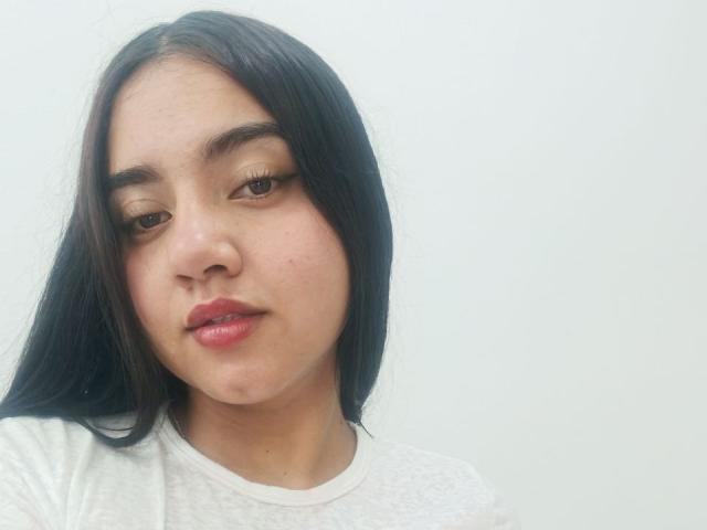 SalomeLee has long black hair, brown eyes, and full pink lips. She's wearing a white shirt, and her skin is slightly flushed. Her expression is neutral, with a slight smile.