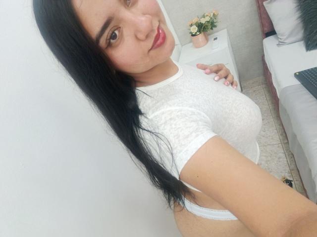 SalomeLee, con i suoi lunghi capelli neri, indossa una camicetta bianca trasparente che mette in mostra il suo seno prosperoso, sorridendo alla fotocamera, in piedi in una stanza con un letto e un piccolo tavolo..