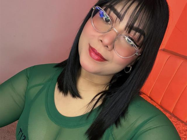 AlexaKiim with black hair, glasses, green sheer top, red lipstick, and hoop earrings, smiling at the camera, showing her tits through the translucent fabric.