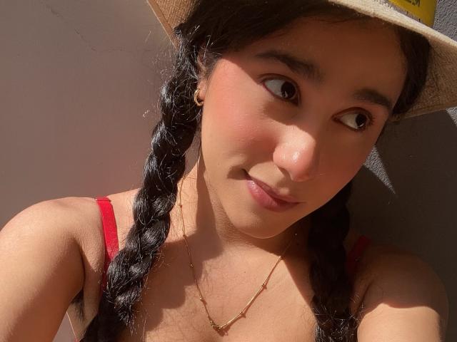TiffannyRose mit geflochtenem schwarzem Haar, die einen Strohhut, ein rotes Kleid mit Trägern, eine goldene Halskette und ein leichtes Lächeln trägt, mit Sonnenschein im Gesicht, süßen braunen Augen, einer kleinen Nase und rosafarbenen Lippen..