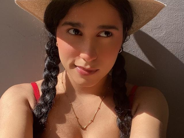 TiffannyRose, met gevlochten zwart haar, een rode tanktop, een gouden ketting en een zonnehoed, heeft een schattig gezicht, kleine borsten en een lichtbruine huid, en kijkt in het zonlicht naar de zijkant..